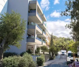Prime urban integration | Modern residential architecture seamlessly blended into the quiet, green landscapes of Galatsi. Exterior 3D architectural rendering showing a side street view of a modern minimalist apartment building in Galatsi, integrated into a green neighborhood next to Veikou Hill.