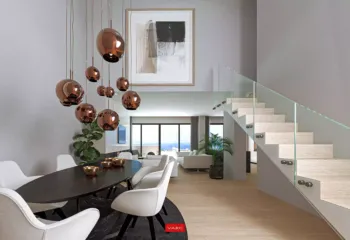 Elegant dining and architectural transitions at the Amerikis 2 duplex penthouse. Interior 3D architectural rendering of a luxury dining area in Ano Glyfada, featuring copper pendant lights, a modern wooden staircase with glass railings, and a double-height ceiling.