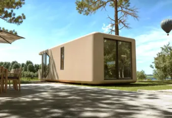 Exterior 3D architectural rendering of a modular capsule home on a sunny day, featuring an outdoor wooden dining set, shadows on the patio, and a hot air balloon in the blue sky.